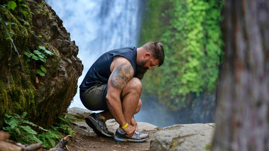 A fit adult male bending down to tighten his shoe laces while on a hike near a waterfall.