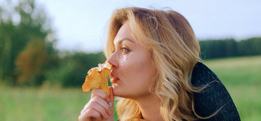 A blonde haired woman standing in a field smelling a mushroom.